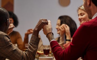 Multi-ethnic group of people holding hands in prayer at Thanksgiving dinner with friends and family, focus on foreground, copy space